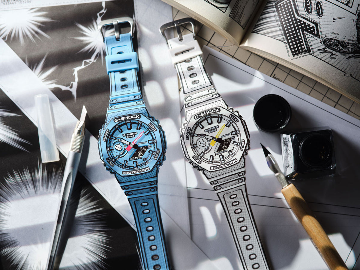 Two G-Shock watches with a 2D manga-style design in blue and white, displayed on a desk with drawing tools and comic pages.