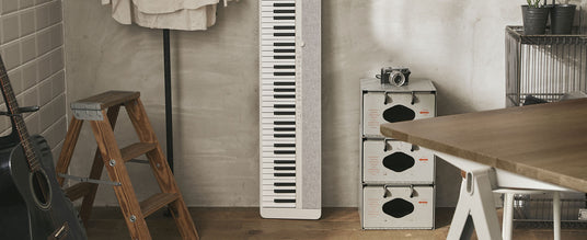 Slim white digital piano standing vertically against a concrete wall next to a wooden ladder and vintage metal storage bins.