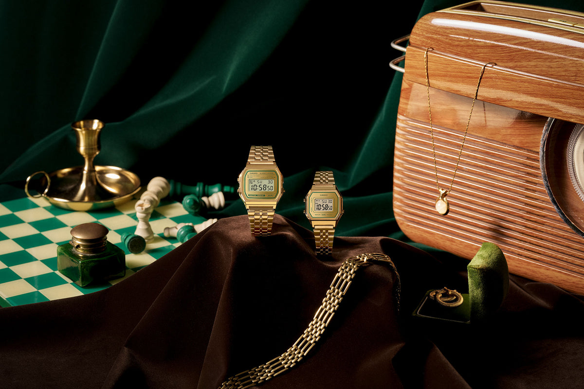 Two gold Casio digital watches displayed on brown fabric with a vintage radio, chess set, and jewelry on a dark green background.