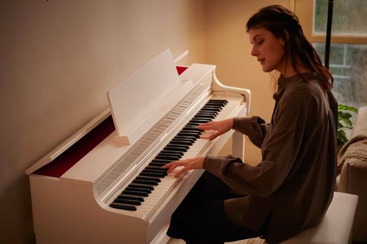 A woman playing a modern white digital piano in a cozy, warmly lit room.