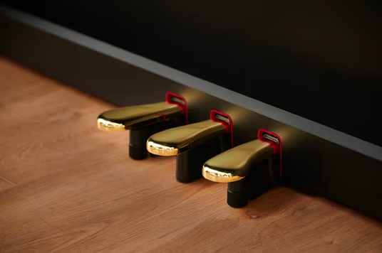Three polished gold piano pedals with red felt accents mounted on a sleek black base above a wooden floor.