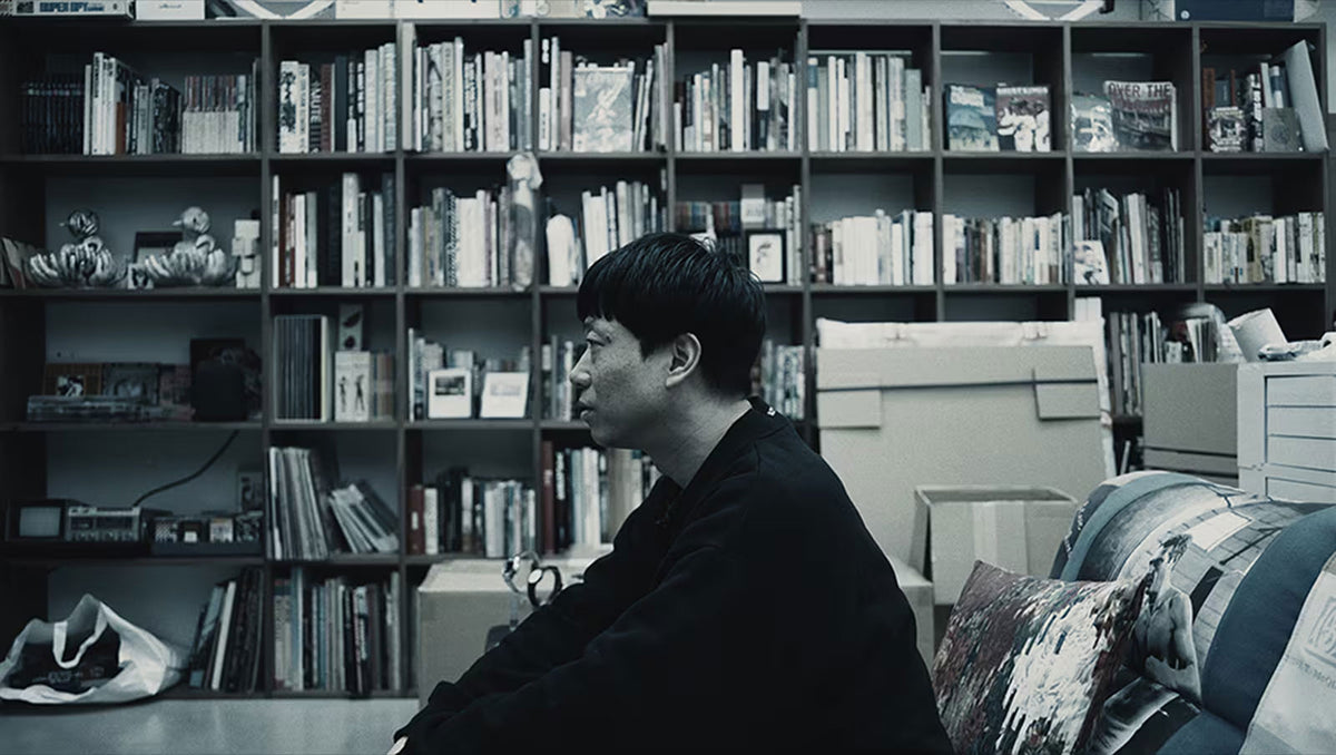Kosuke Kawamura sitting in profile before a large, filled wooden bookshelf in a muted, desaturated studio setting.