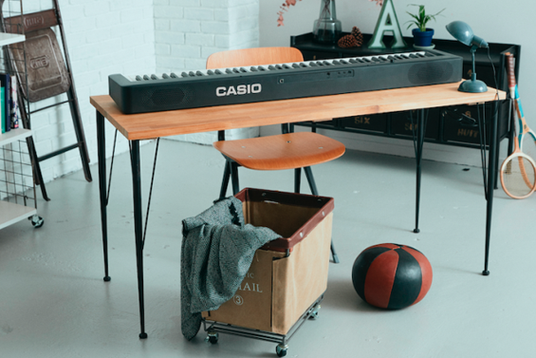 Black Casio digital keyboard placed on a wooden desk with metal legs in a bright, modern studio space.