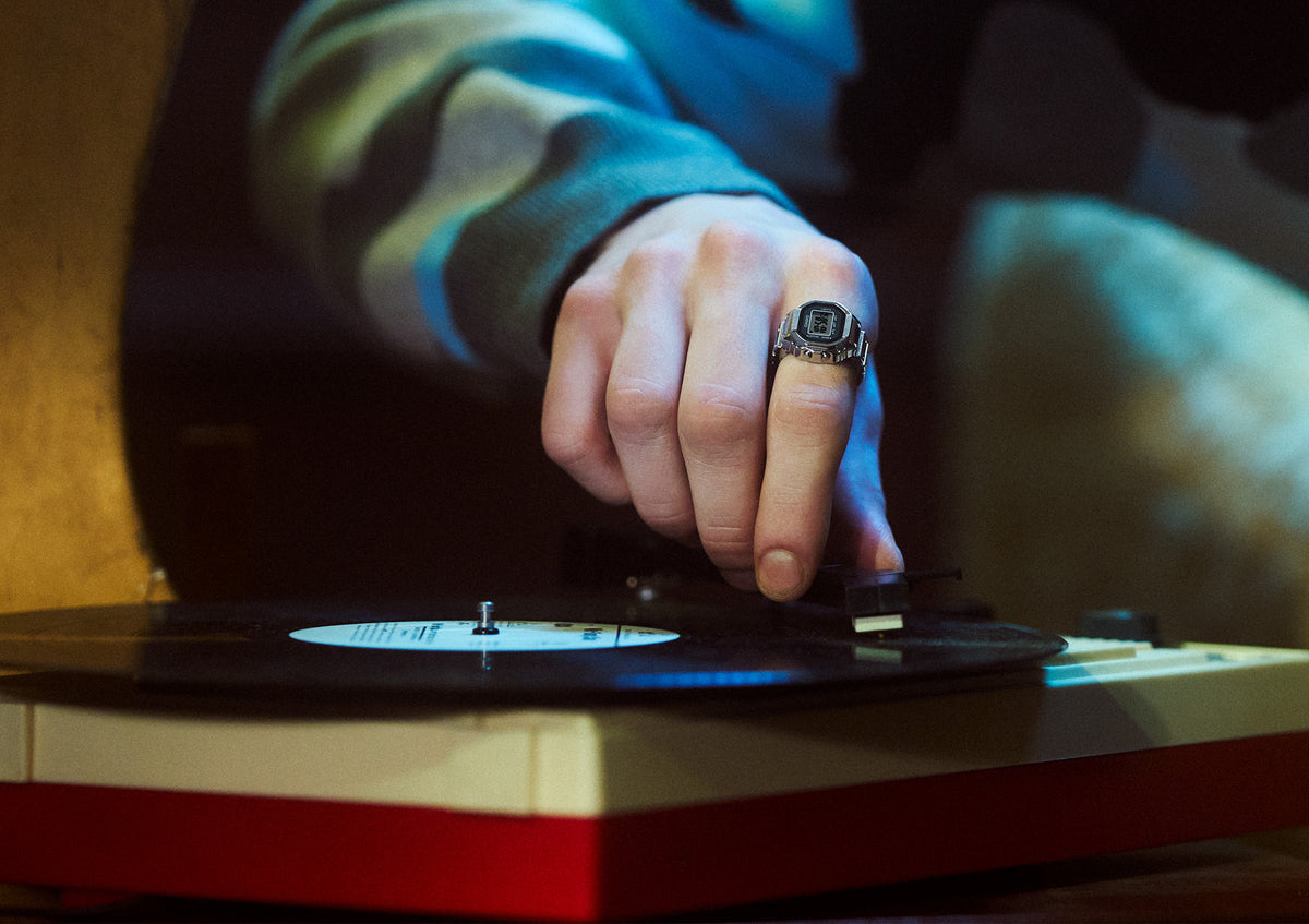 A person wearing a silver Casio digital ring watch places the needle on a black vinyl record spinning on a turntable.