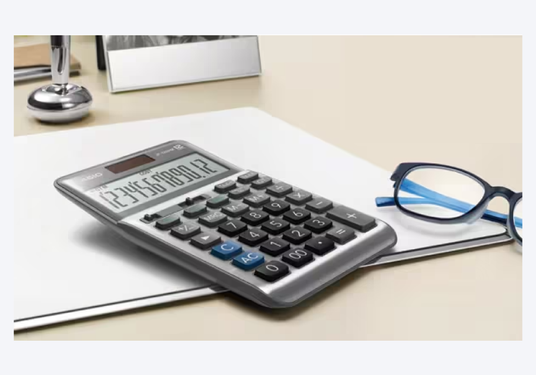 Silver Casio desktop calculator with large display and black keys sitting on a white desk next to blue reading glasses.