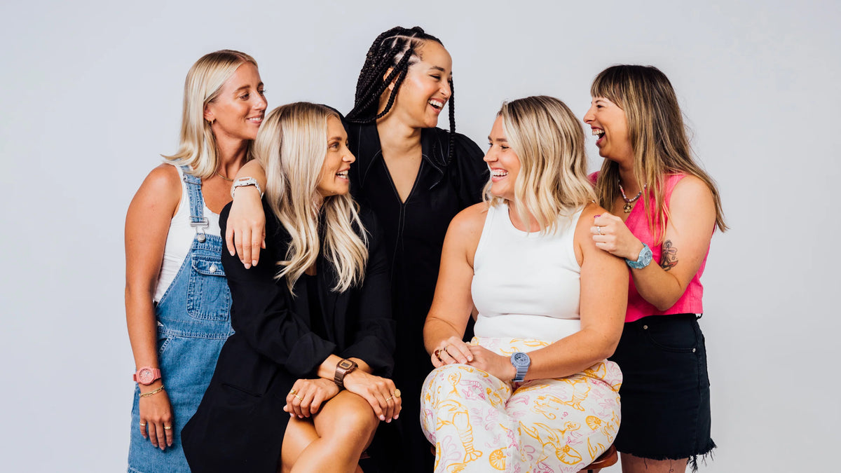 Five diverse women laughing together while wearing casual, colorful outfits against a plain light gray background.