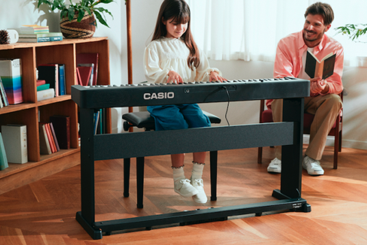 Young girl playing a black Casio digital keyboard on a stand while a man watches in a cozy, sunlit living room.
