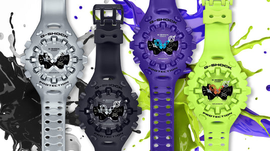 Four G-Shock watches in silver, black, purple, and neon green arranged against a vibrant, multi-colored paint splash.