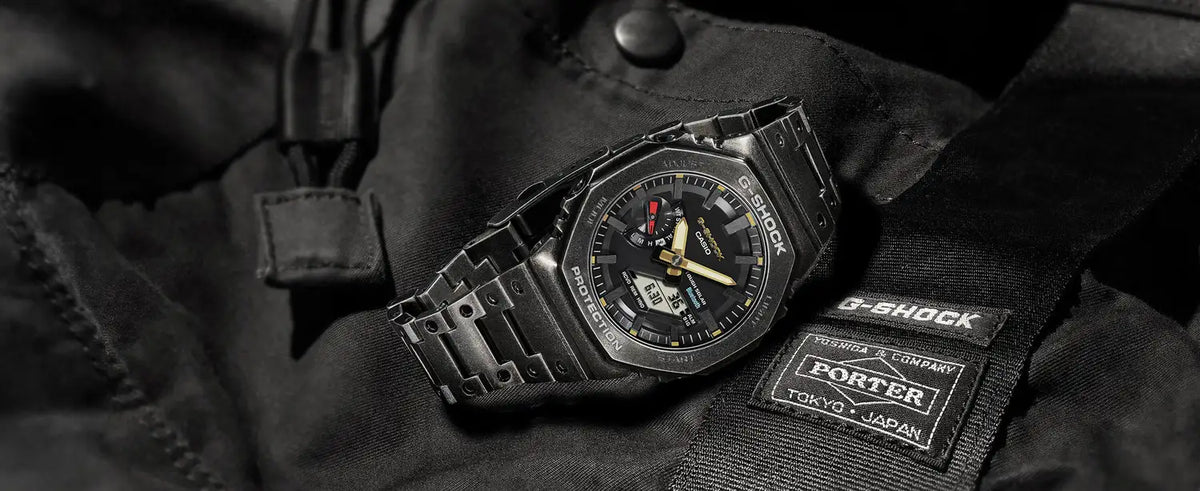 Black metal G-Shock watch with gold accents resting on a black Porter Yoshida & Company branded fabric bag.