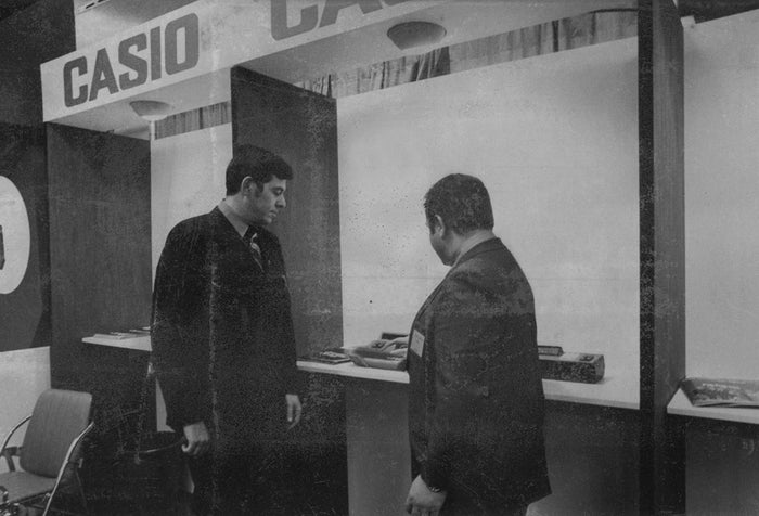 Two men in suits examine vintage Casio electronic calculators at a 1970s trade show booth.