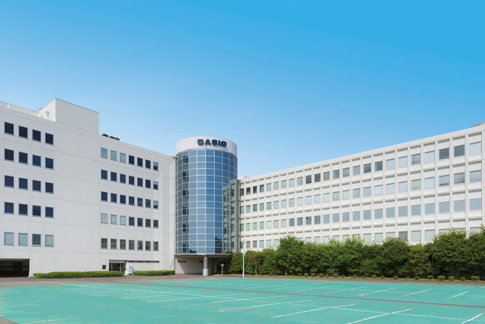 Modern white Casio office building with a central glass cylindrical tower under a clear blue sky.