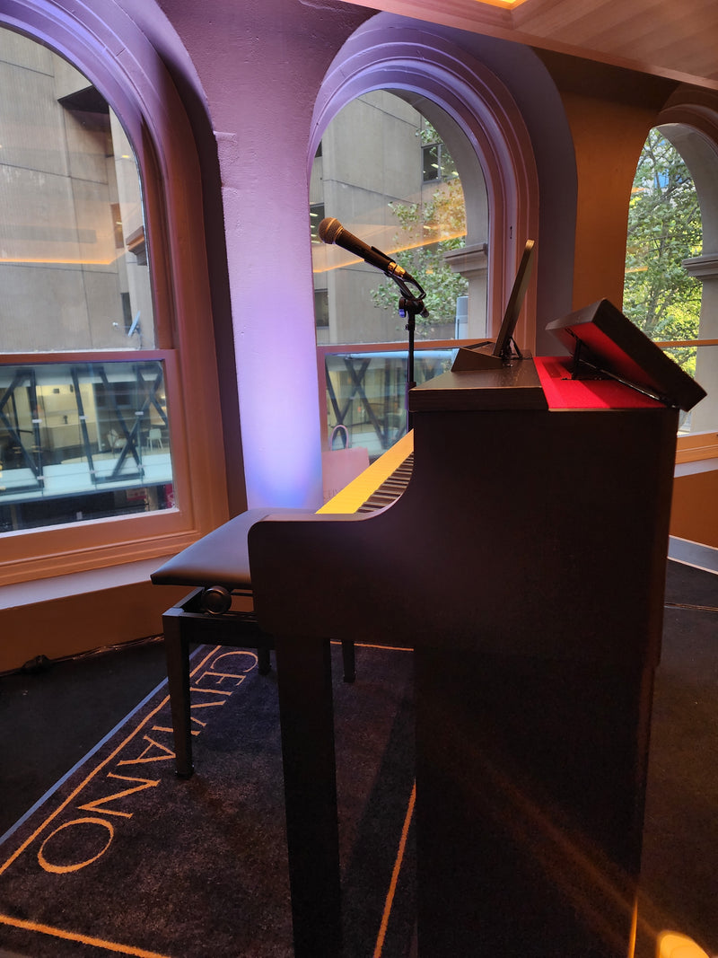 Black digital piano with a microphone stand positioned before arched windows in a room with purple accent lighting.