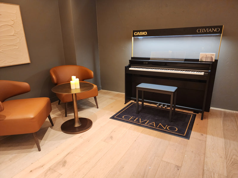 Black Casio Celviano digital piano with bench, two tan leather chairs, and a small table with candles on light wood floors.
