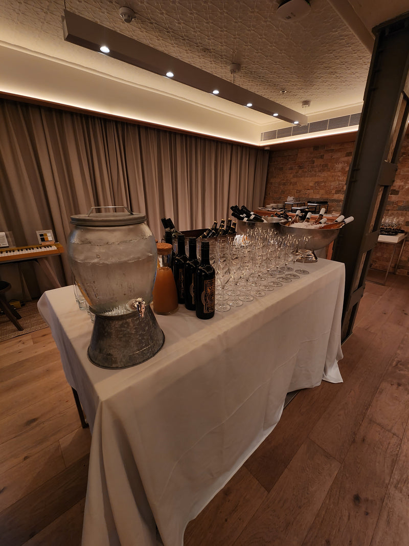White-clothed table featuring a glass water dispenser, wine bottles, and rows of empty stemware in a warm, modern event space.