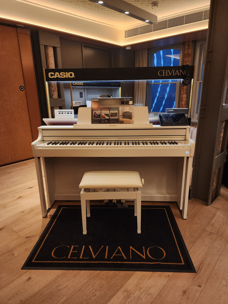 White Casio Celviano digital piano with a matching bench on a branded black floor mat in a modern showroom.