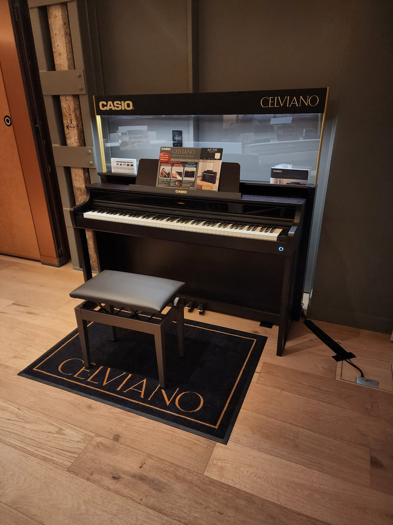 Black Casio Celviano digital piano with matching bench on a branded rug in a modern showroom setting.
