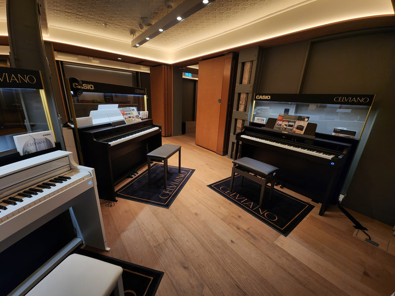 Two black Casio Celviano digital pianos with matching benches on branded floor mats in a modern showroom.