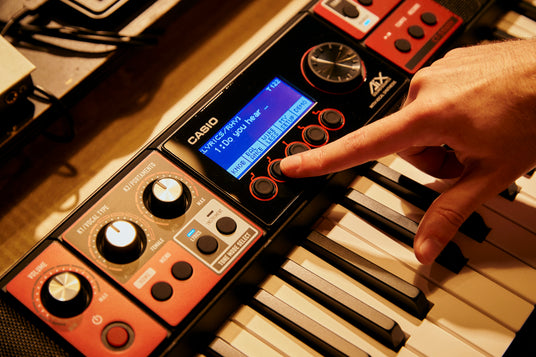 A hand pressing a button on a Casio CT-S1000V keyboard with a glowing blue digital display and red control panel.