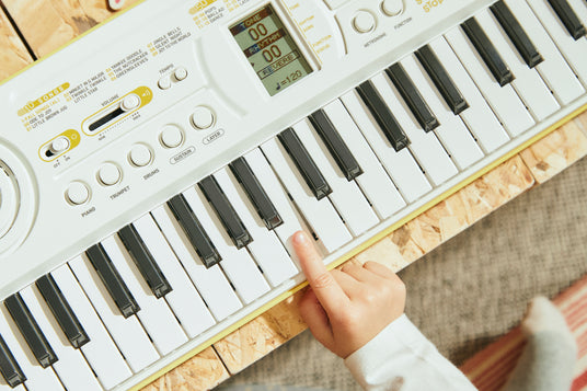 Child's finger pressing a white key on a white electronic keyboard with a digital display and song list.