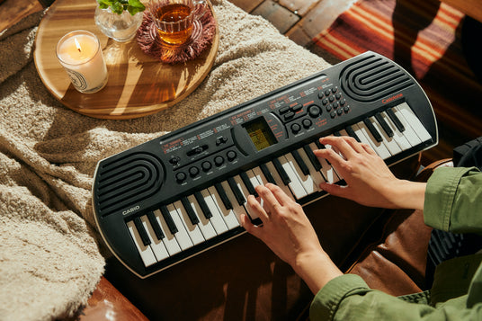 Hands playing a black Casio SA-81 portable keyboard on a brown leather sofa next to a candle and tea.
