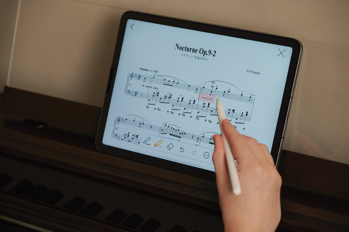Hand using a stylus to edit Chopin sheet music on a tablet screen resting on a brown digital piano.