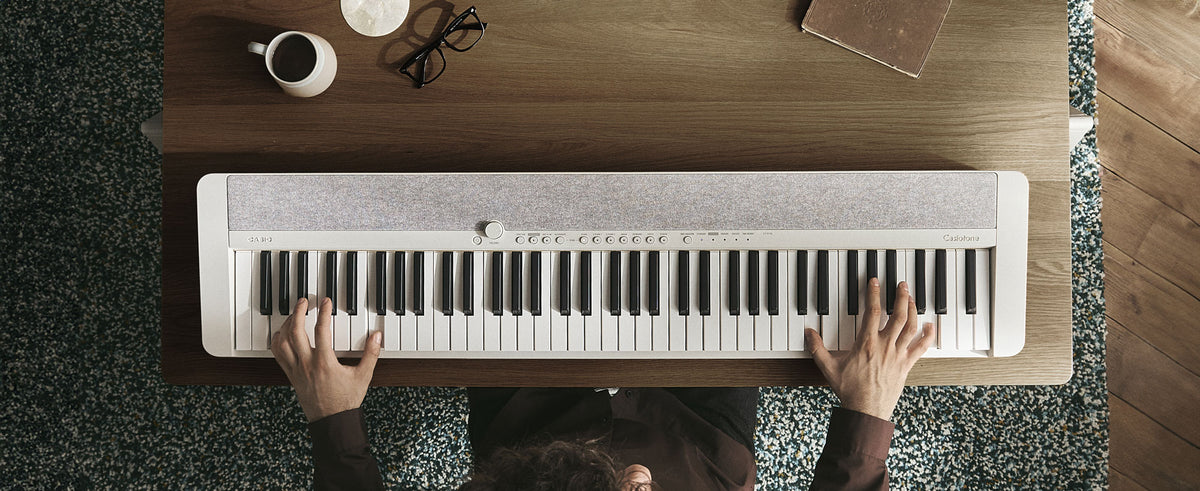 Top-down view of a person playing a sleek white Casio Casiotone keyboard on a wooden desk with a coffee mug and glasses.