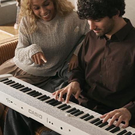 A man playing a white Casio digital piano while a woman points at the keys, sitting together on a cozy couch.