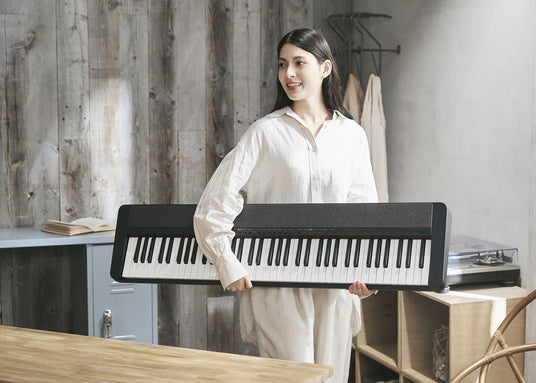 Smiling woman in a white linen shirt holding a sleek black digital piano in a rustic, modern studio setting.