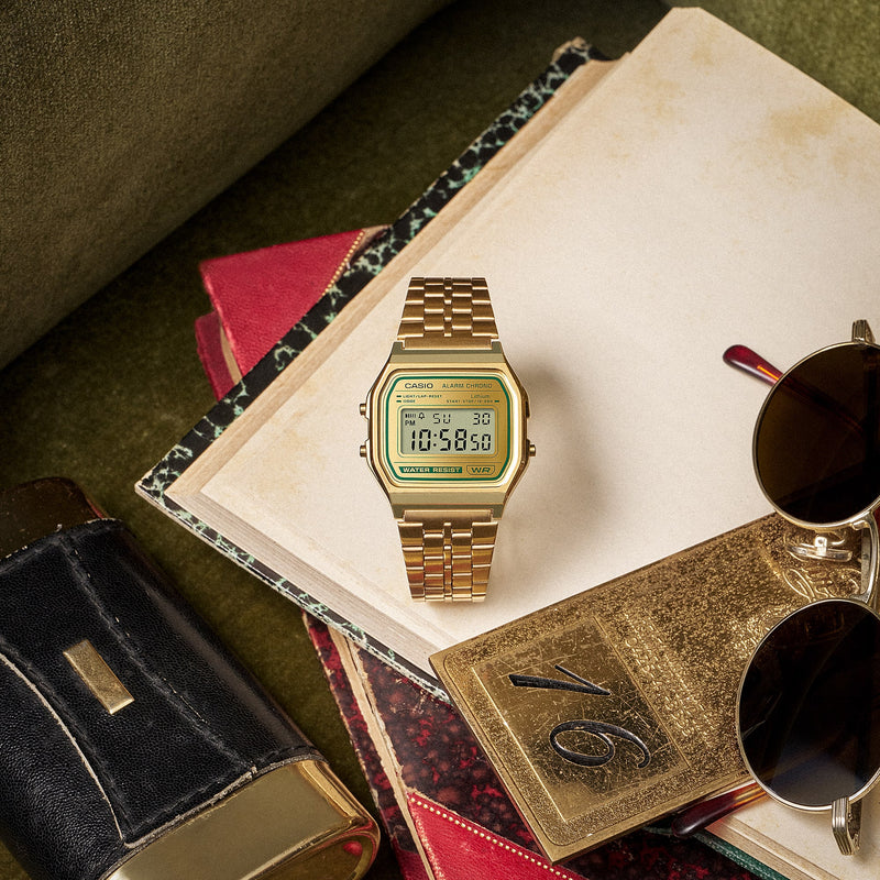 Gold-tone Casio digital watch with a green-accented face resting on vintage books and sunglasses.
