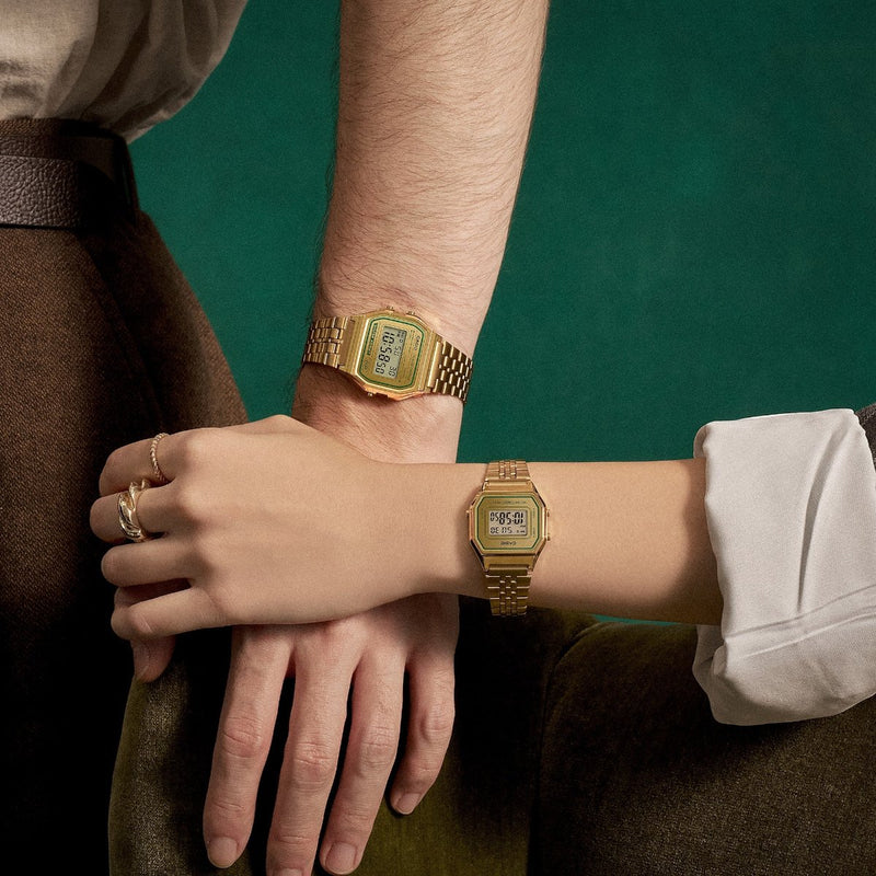 Two people wearing matching gold-tone Casio digital watches with metal link bracelets against a dark green background.