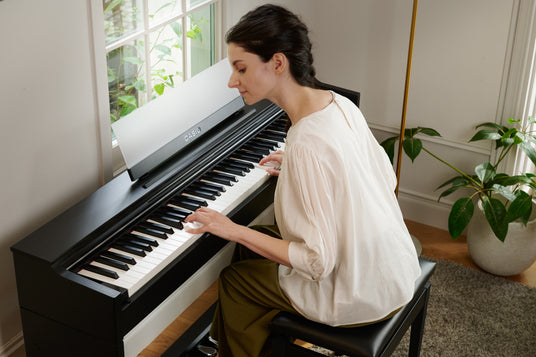 Woman playing a sleek black Casio digital piano in a bright, modern living room near a window.