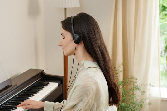 Woman with long dark hair wearing black headphones while playing a dark wood digital piano in a bright, cozy room.