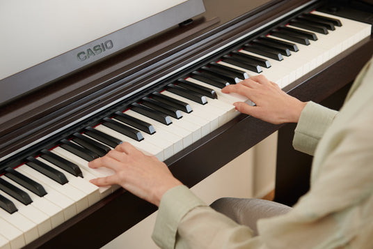 Hands playing a dark wood Casio digital piano with white and black keys in a bright, modern room.