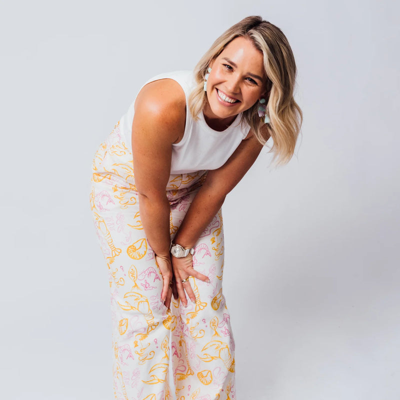 Blonde woman smiling while wearing a white tank top and wide-leg pants with a yellow and pink lobster print.