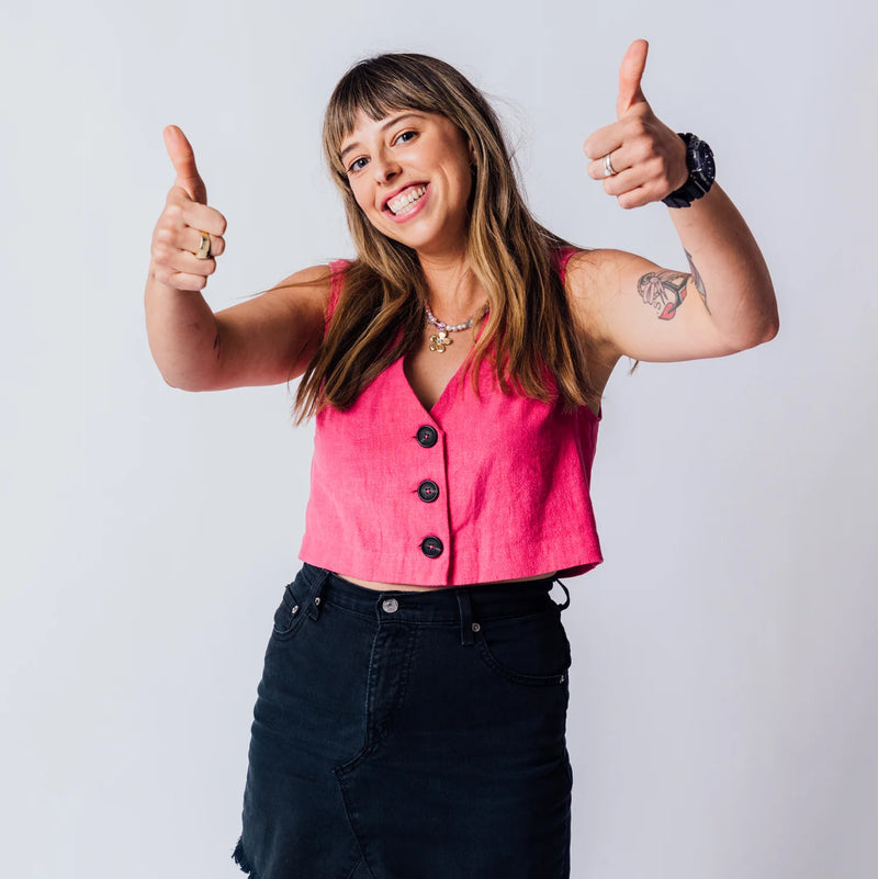Smiling woman with bangs wearing a pink button-down vest and black skirt giving a double thumbs up against a white background.