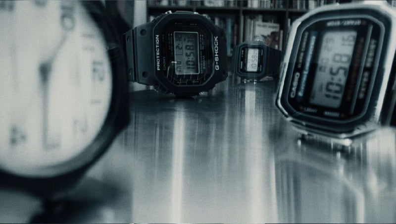 Three Casio digital watches arranged on a reflective surface with a blurred analog clock in the foreground.
