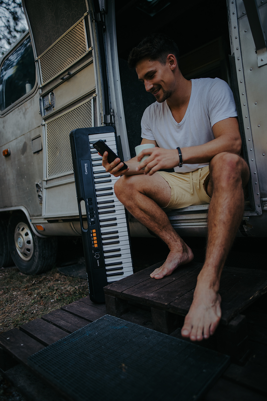 Man sitting in a vintage camper doorway holding a phone and mug next to a black Casiotone keyboard.