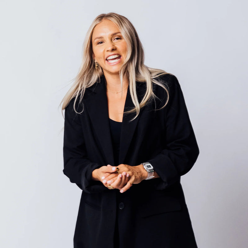 Blonde woman laughing while wearing a black blazer over a matching top against a plain light gray background.