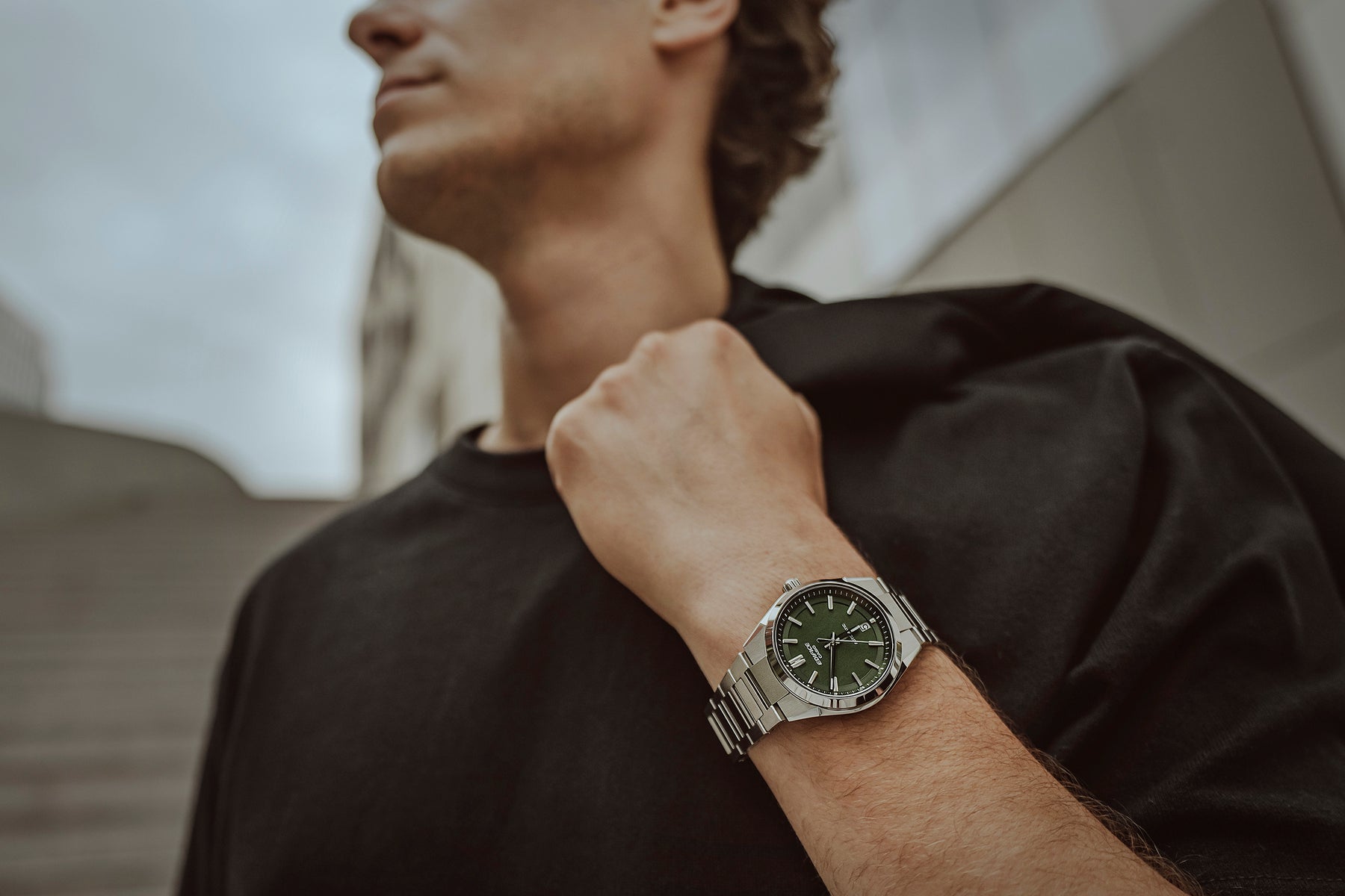 A man wearing a silver-toned metal watch with a dark green dial and matching green accents, shown in a lifestyle outdoor setting.