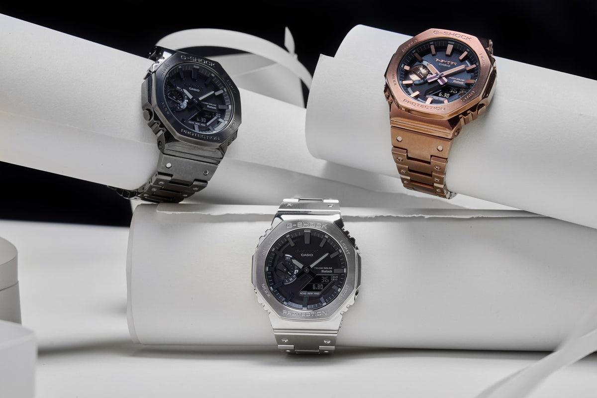 Three G-Shock watches in silver, black, and rose gold metal finishes displayed on white cylindrical pedestals.