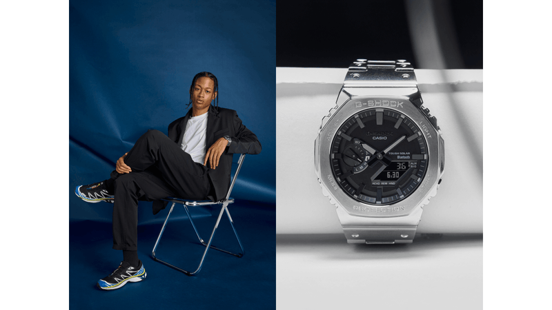 Man in a black suit sitting on a chair next to a stainless steel G-Shock GMCB2100D-4 watch with a black analog-digital dial.