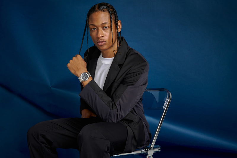Young man in a black blazer wearing a silver G-Shock GMCB2100D-9 watch, seated against a deep blue background.