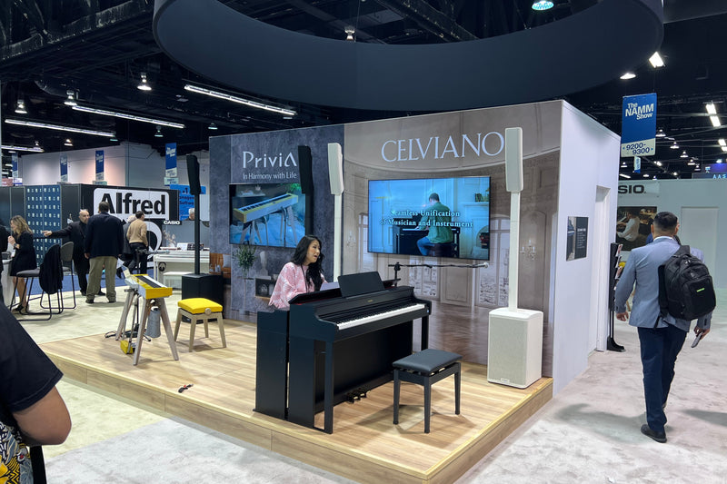 Woman playing a black Casio Celviano digital piano at a trade show booth with screens displaying product information.