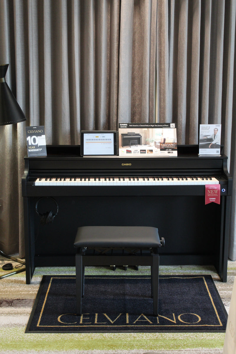 Black Casio Celviano digital piano with a matching bench and tablet on a branded rug in front of grey curtains.