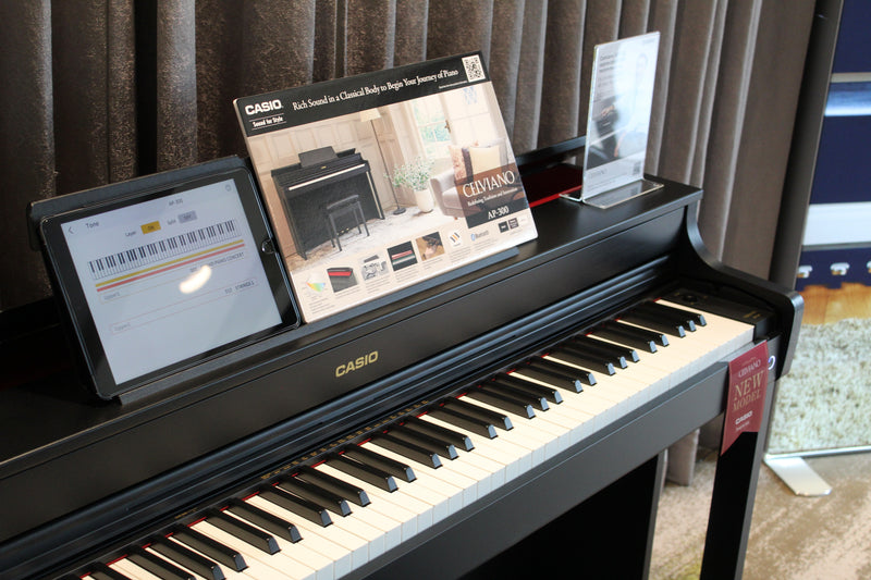 Black Casio Celviano AP-300 digital piano with a tablet display and promotional signage on a stand.