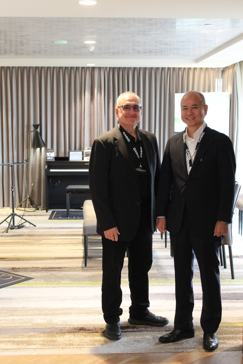 Two men in formal black suits and Casio lanyards standing in a modern hotel room with a piano in the background.