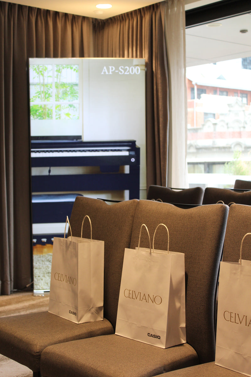 White Celviano Casio gift bags placed on grey upholstered chairs in front of a Casio AP-S200 digital piano display banner.