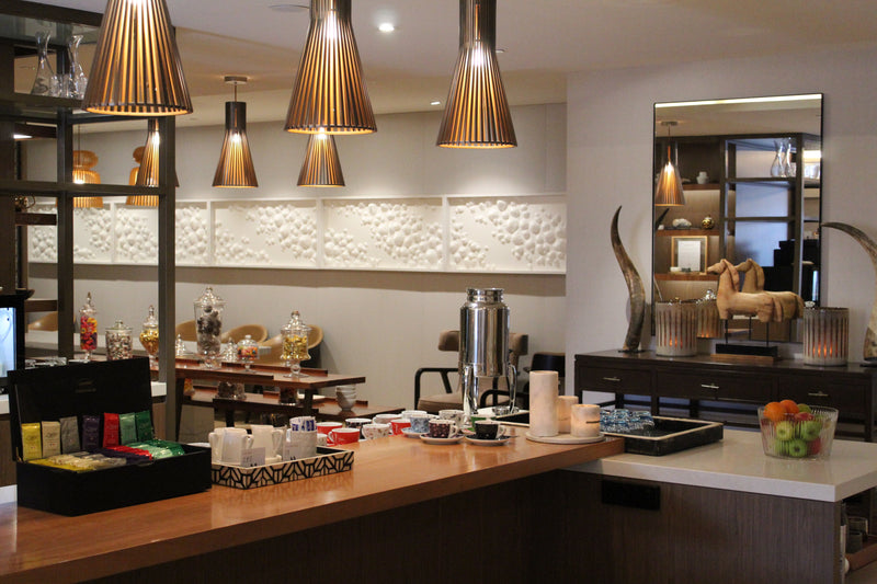 Modern hotel lounge with wooden counters, pendant lights, tea selection, and decorative wall art in a warm, elegant setting.