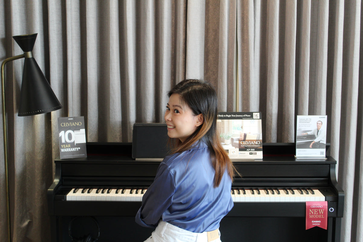 Smiling woman in a blue shirt sitting at a black Casio Celviano digital piano in front of grey curtains.