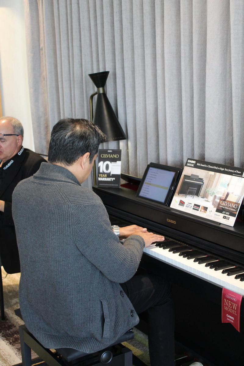 Man in a gray sweater playing a black Casio Celviano digital piano with a tablet and warranty sign nearby.